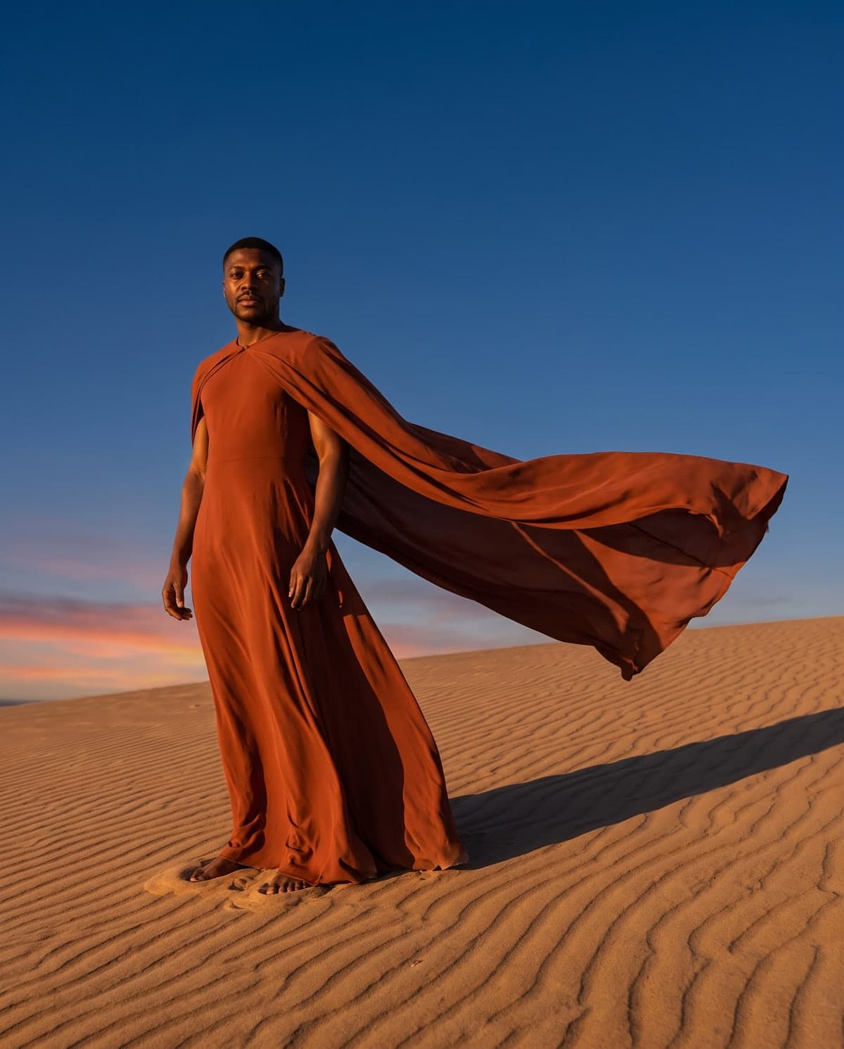 AI desert fashion editorial of a man in a flowing rust outfit on a sand dune at sunset