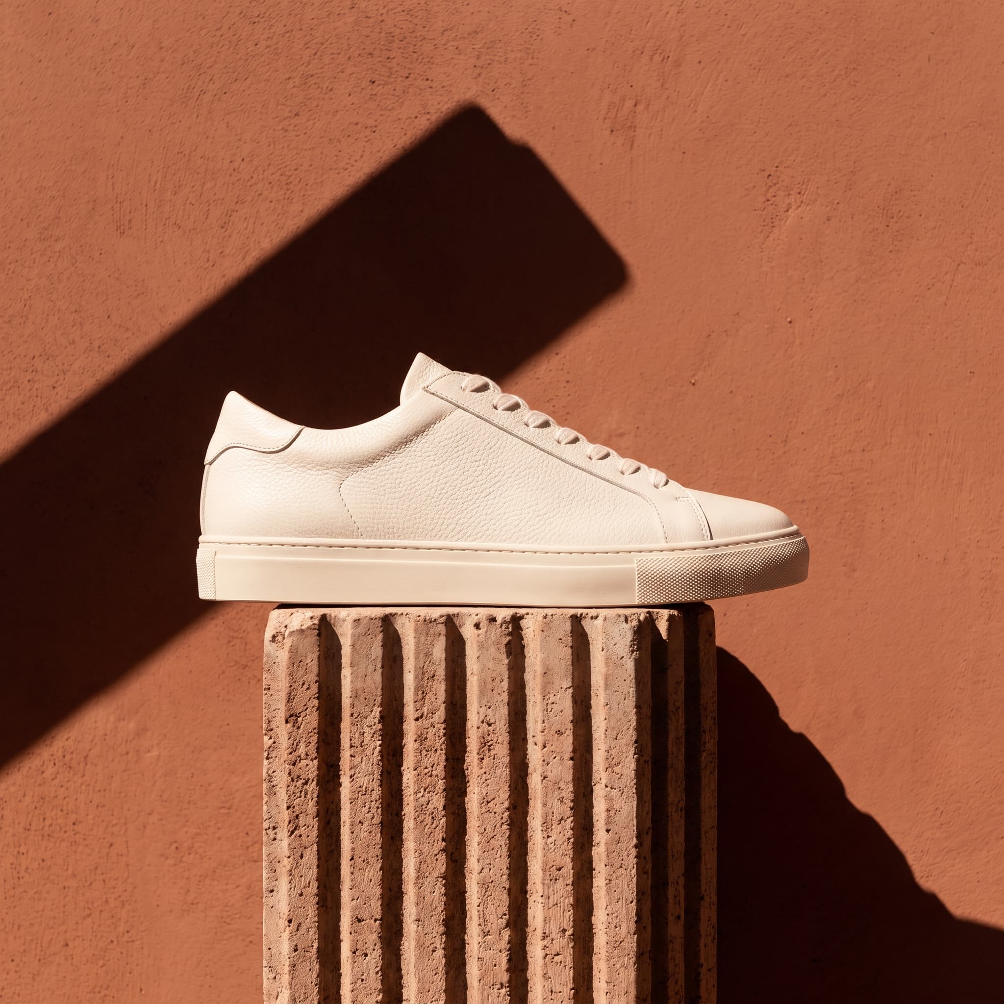 Plain white sneaker on a ribbed concrete block against a terracotta wall in hard sunlight
